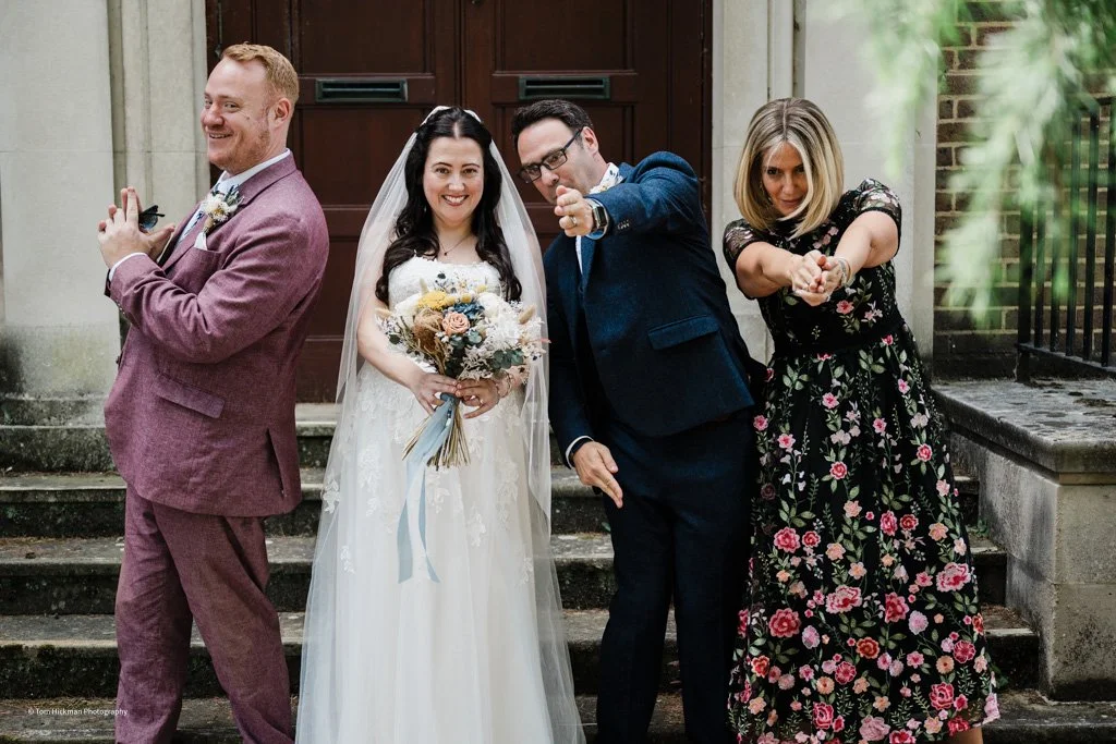 Four people, a bride, groom, man, and woman, posing playfully outside a building with steps, smiling and pointing at the camera.