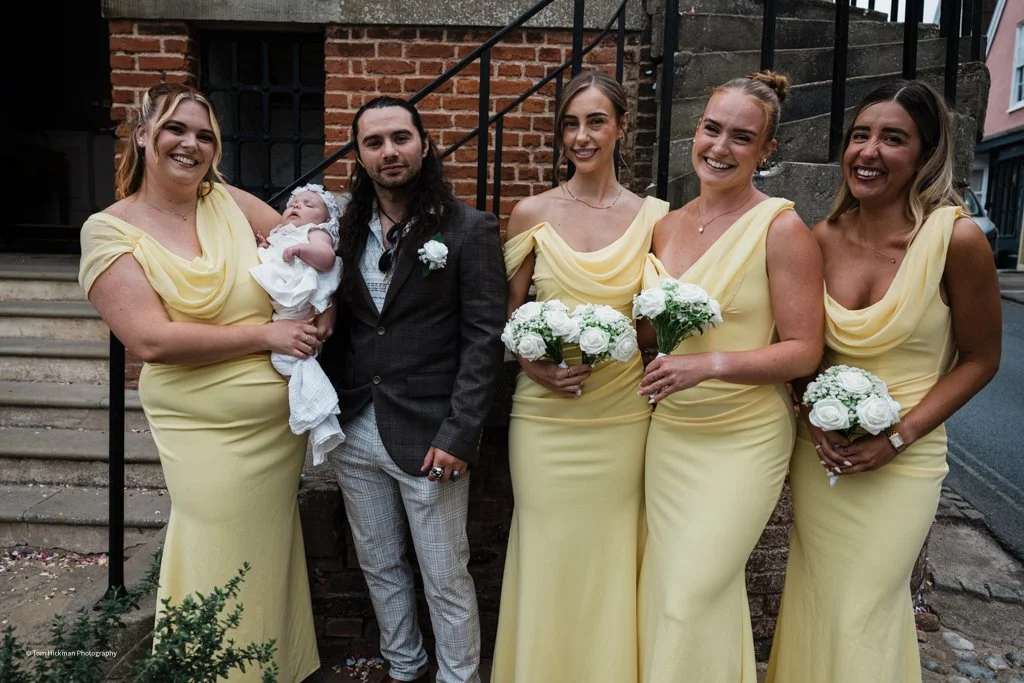 Group of five women and one man standing outdoors, five women in yellow dresses holding bouquets, one woman holding a baby, brick building in background.