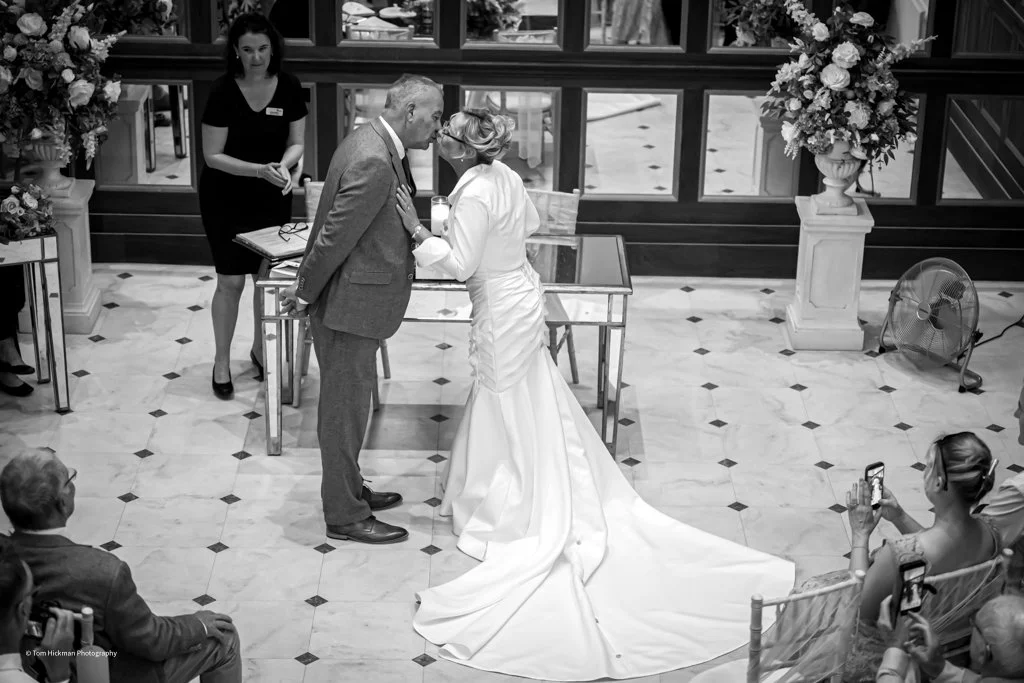 A wedding ceremony with a couple kissing, surrounded by guests taking photos, with floral arrangements and a marble floor.