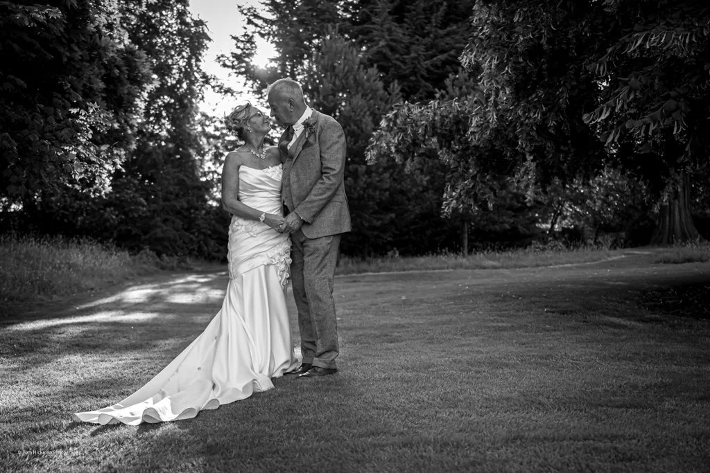 Black and white photo of a bride and groom standing close together outdoors, holding hands, with trees in the background.