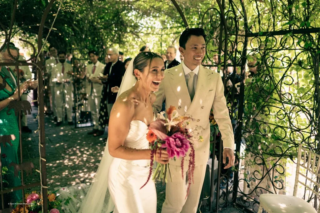 Bride and groom smiling as they walk through a garden archway, surrounded by onlookers at a wedding ceremony.
