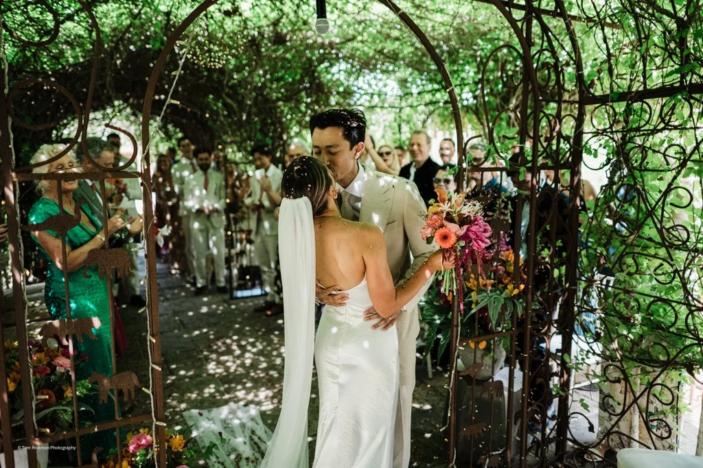 A bride and groom share a kiss during their outdoor wedding under a decorative metal arch, surrounded by friends and family, with green foliage overhead.