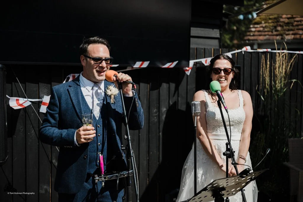 A man and a woman dressed in formal attire, standing outdoors with microphones, likely at a wedding reception, smiling and wearing sunglasses.
