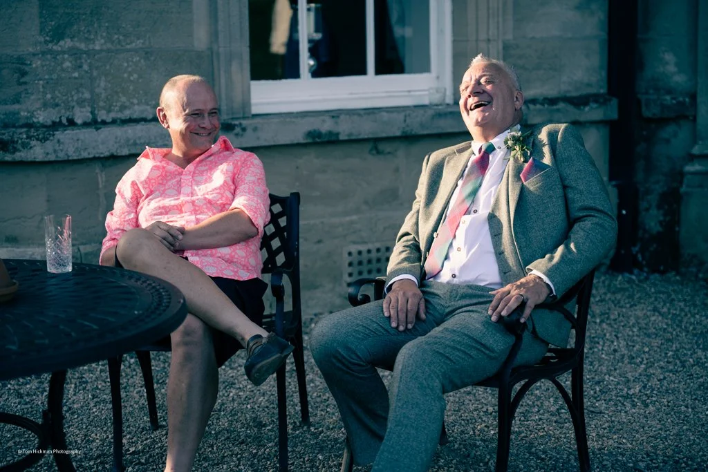 Two men sitting outdoors, laughing and enjoying each other's company, one wearing a pink shirt and the other in a gray suit with a boutonniere.
