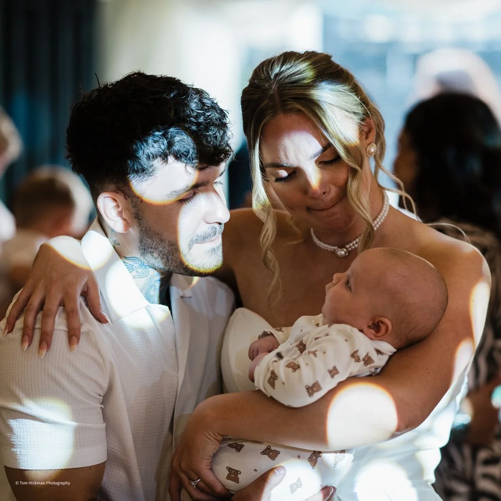 A woman holding a baby while a man looks on with a serious expression during a special occasion, likely a christening or baptism.