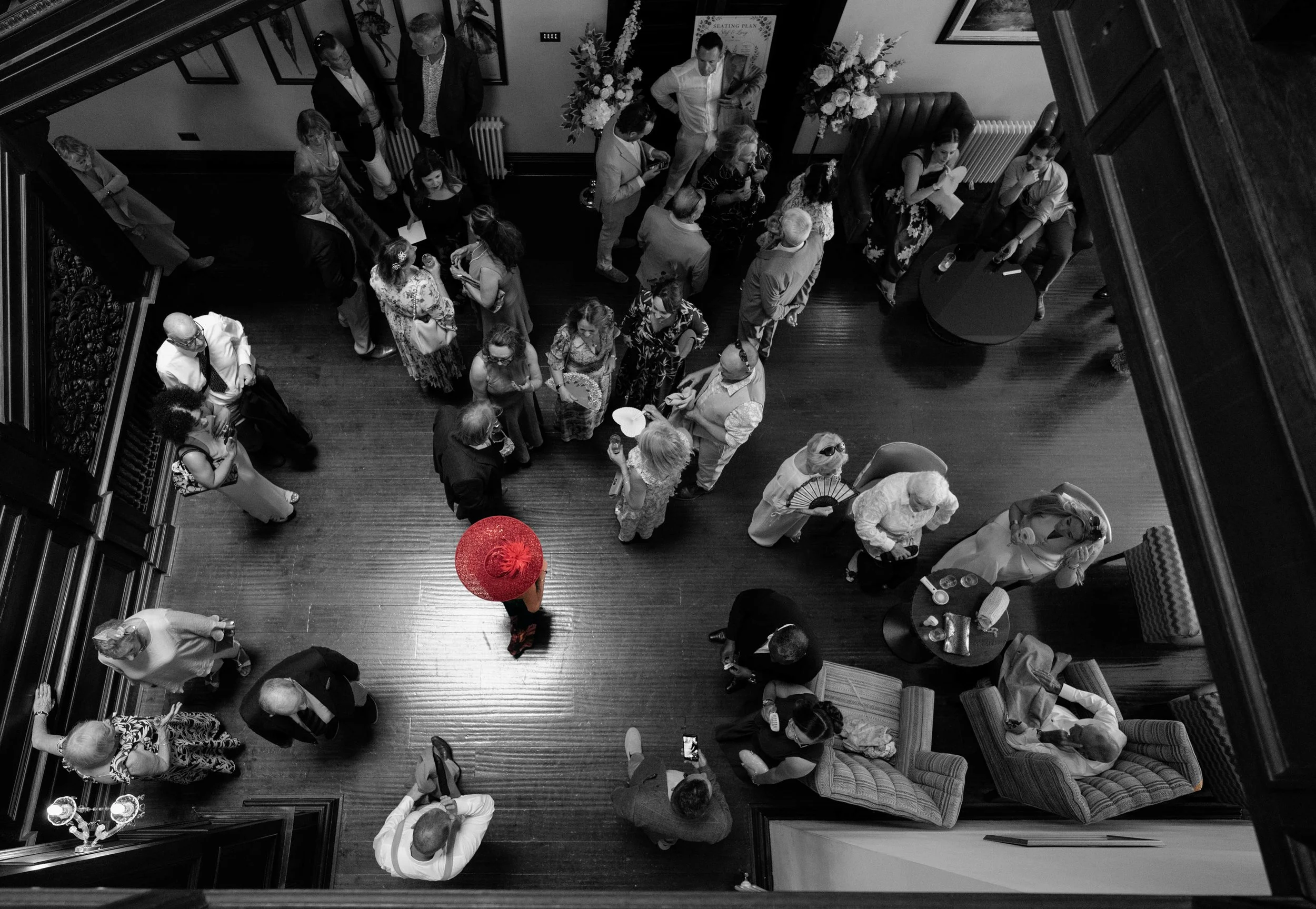 People gathered in a room for a social event, viewed from above, with floral arrangements and a seating plan in the background.