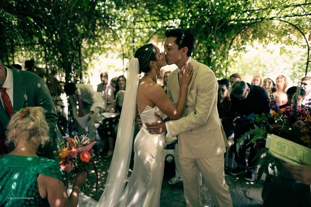 Couple shares a kiss during their wedding ceremony outdoors, surrounded by seated guests in shaded greenery.