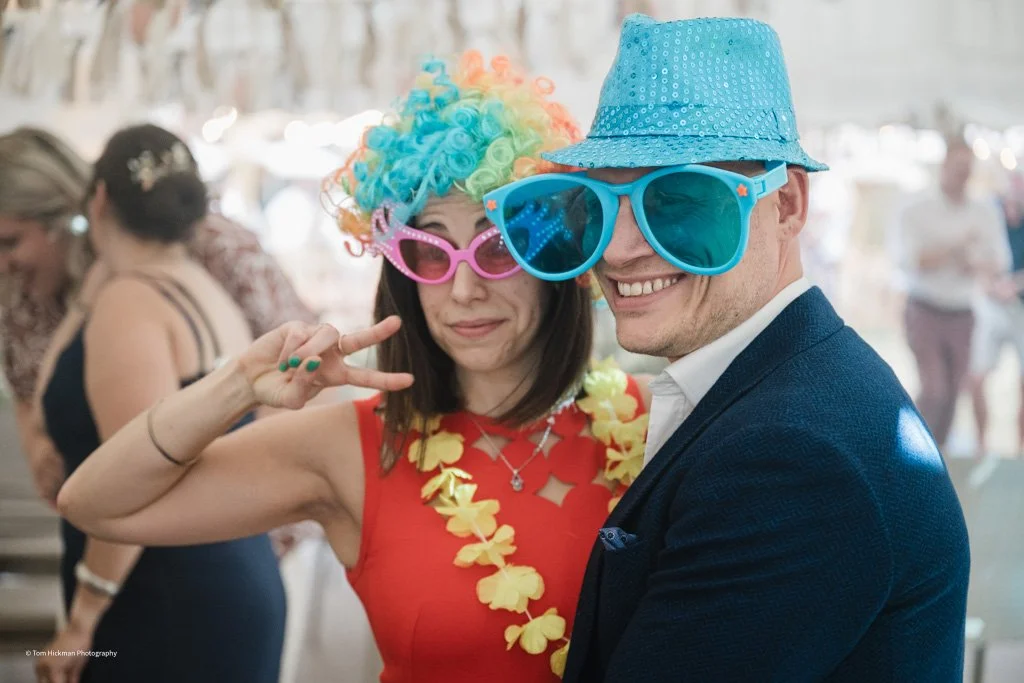 A woman and a man dressed up with large sunglasses and colorful accessories at a party. The woman wears pink sunglasses, a rainbow wig, and a red dress with a yellow floral lei, making a peace sign with her fingers. The man wears blue sunglasses, a b