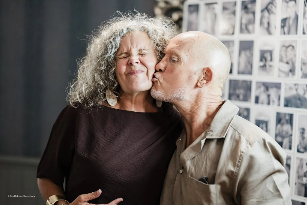 A woman with curly gray hair and closed eyes receives a kiss on the cheek from a bald man with a gray beard in an indoor setting with black and white photos on the wall.