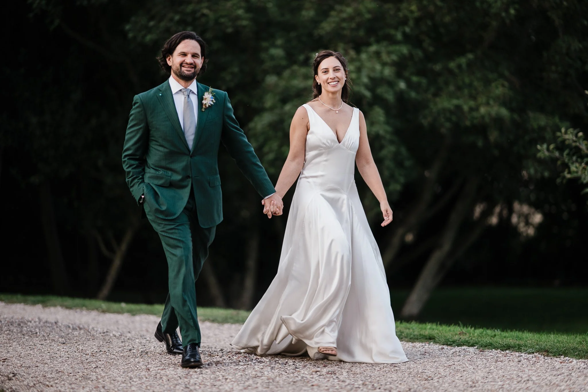 A bride and groom holding hands and walking outdoors, with the groom in a green suit and the bride in a white wedding dress, on a gravel path surrounded by trees.