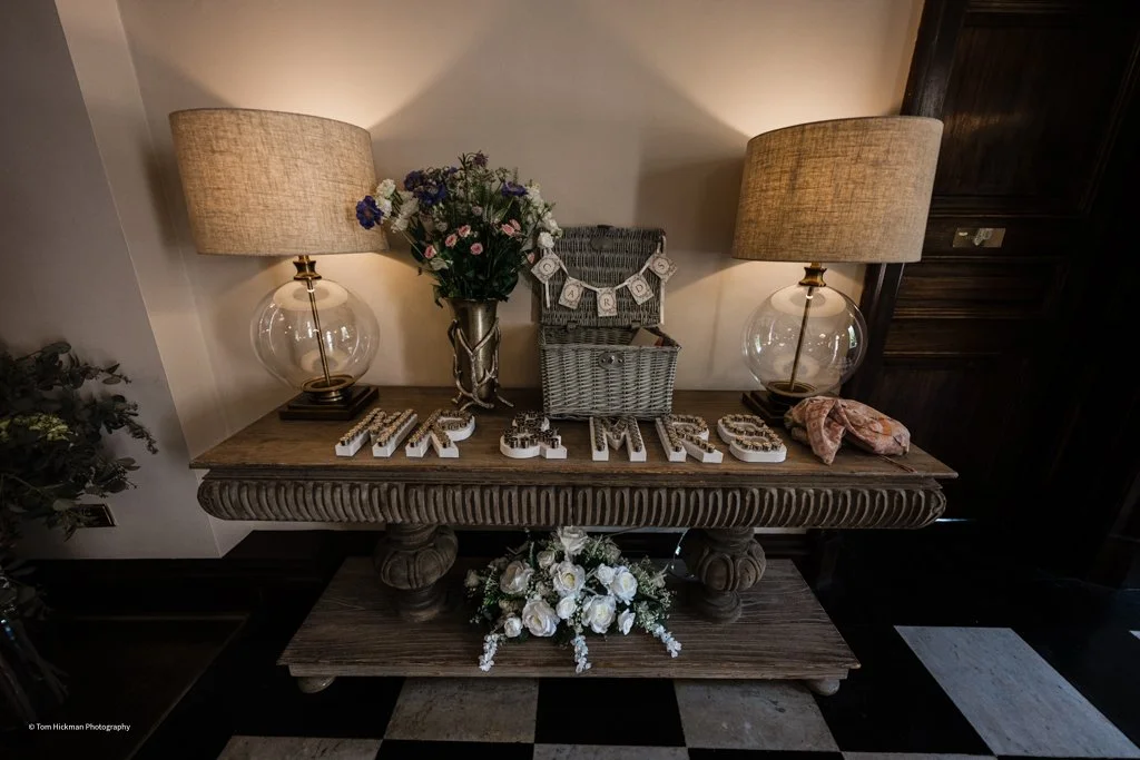 A wooden table decorated with a bouquet of flowers, two lamps, a small Lego garland, a wicker box, and a pink cloth, with a sign spelling out 'Mr & Mrs' on the table and a floral arrangement underneath.