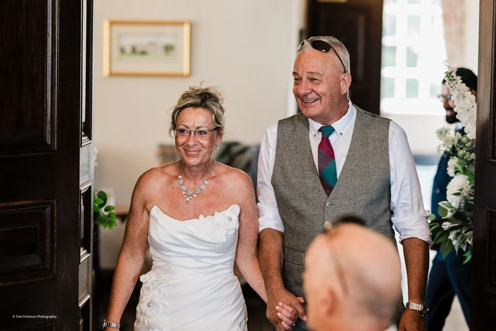 A smiling woman in a white wedding dress holding hands with a man in a gray vest, tie, and white shirt, entering a room decorated with flowers and framed pictures.