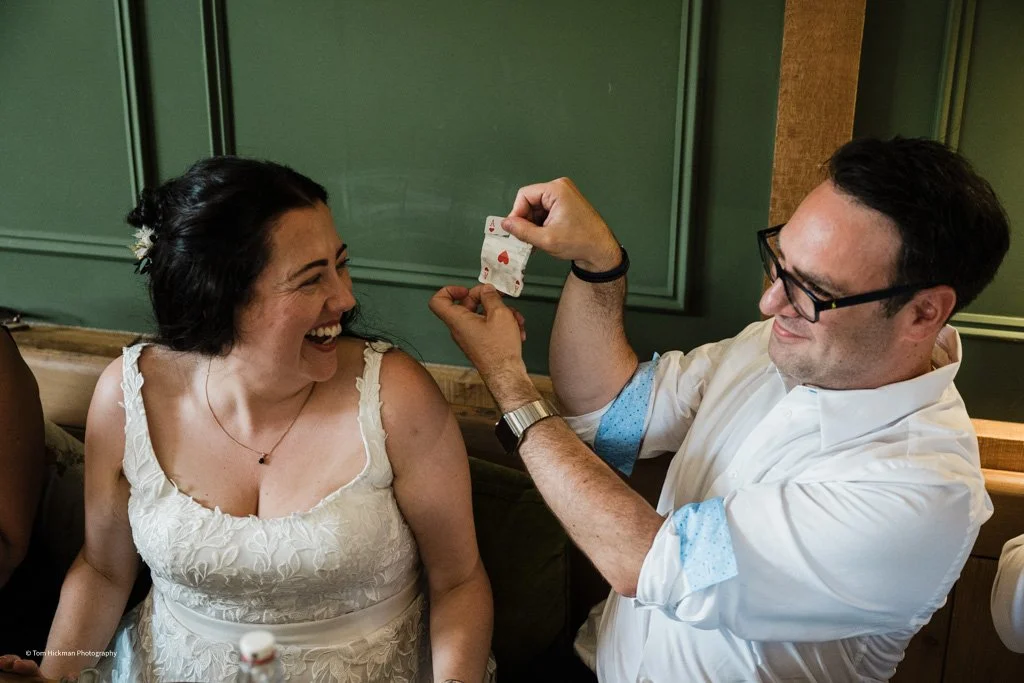 A man with glasses and a white shirt secretly holds two playing cards behind his back, showing a six and a four of hearts, while a woman in a white dress laughs and looks at him.