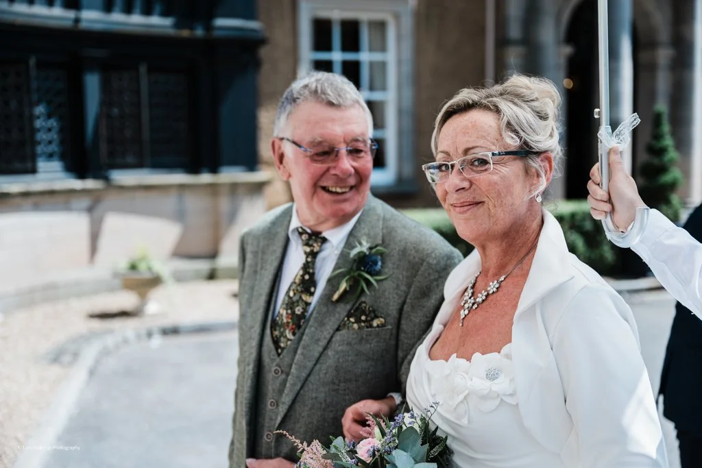 An elderly couple at a wedding, smiling and holding hands outdoors, with a woman in white holding a bouquet of flowers beside them.