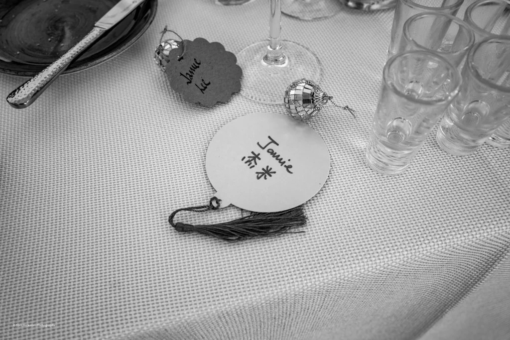 A table setting with a name tag labeled 'Jamie' and decorated with a tassel, a small disco ball, and a list of water glasses on a textured tablecloth.