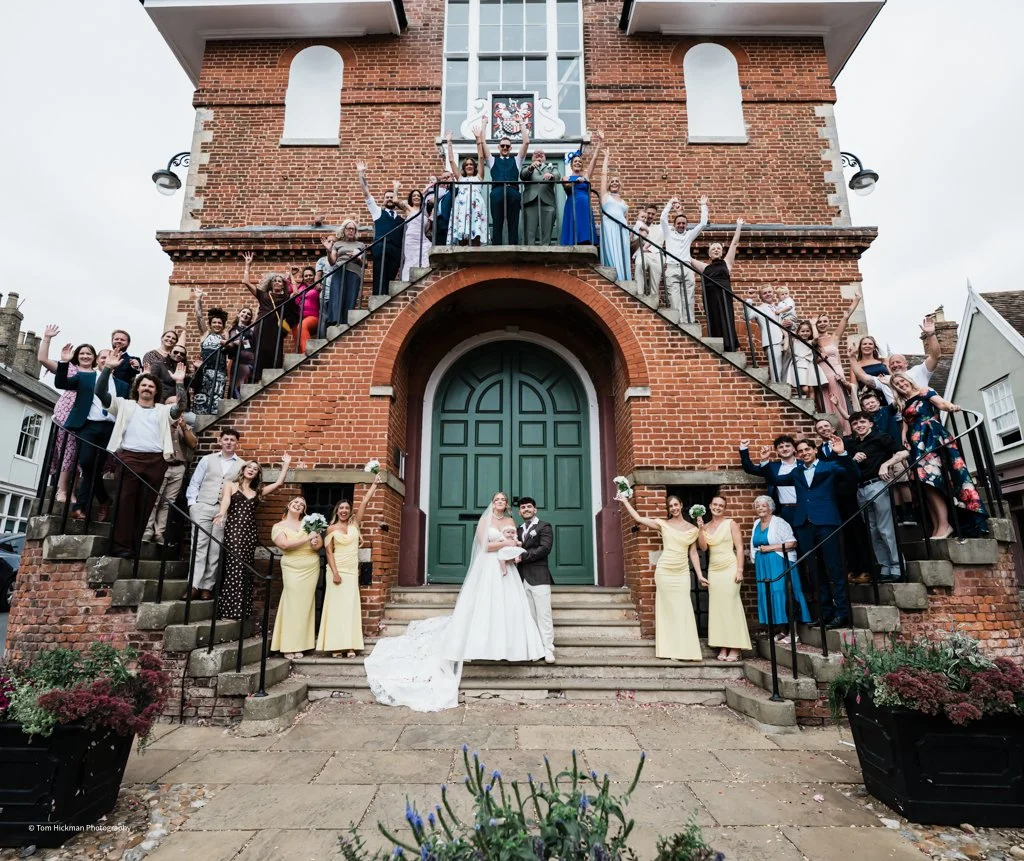 A large group of wedding guests and the bride and groom posing on the steps of a brick building, celebrating with raised hands and smiles.
