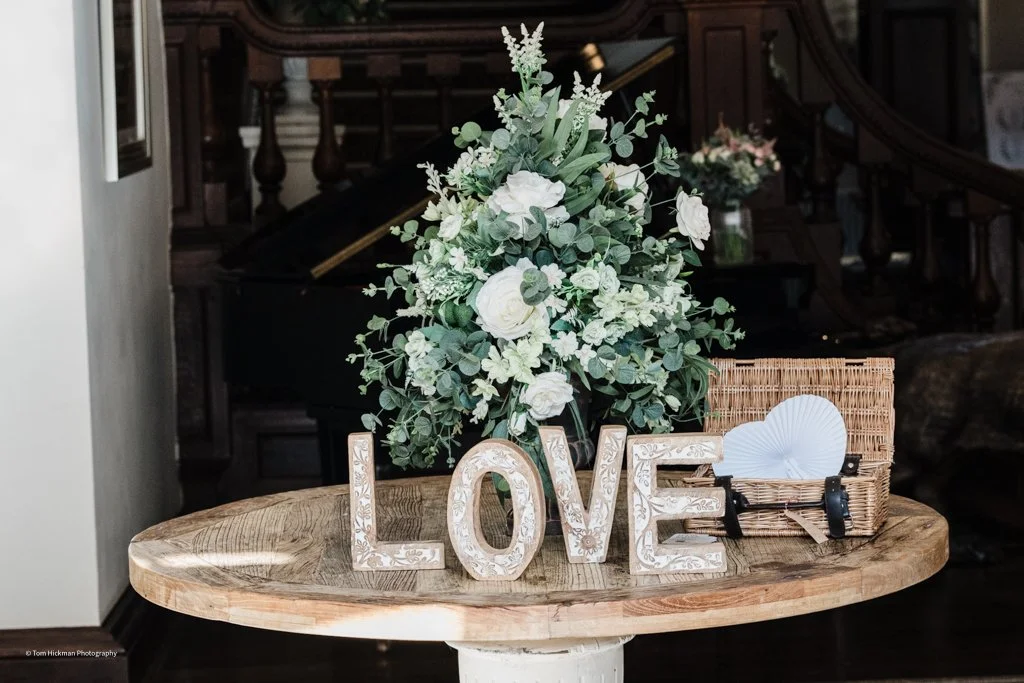A decorative arrangement on a wooden table featuring a large bouquet of white and green flowers, the word LOVE in large white ornate letters, and a small wicker basket holding paper fans.