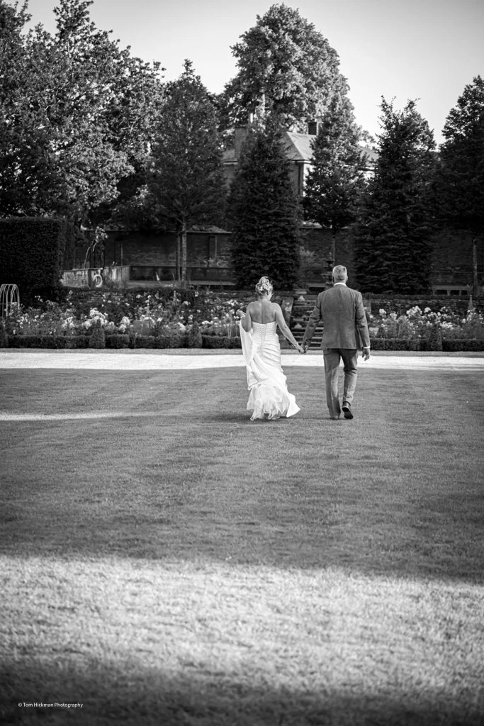 Black and white photo of a bride and groom walking hand in hand on a grassy lawn in a garden or park area with trees and a house or building in the background.