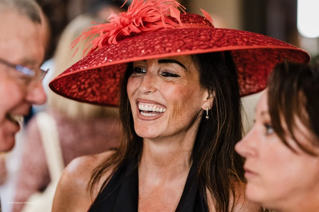 Woman wearing a large red hat with feather decorations, smiling and laughing at a social event.