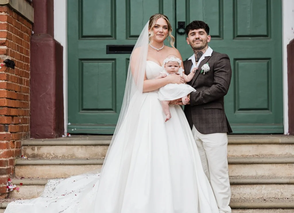 A bride and groom standing on steps outside a building, holding a baby girl. The bride is wearing a white wedding gown and veil, and the groom is wearing a dark suit jacket, white pants, and a boutonniere. The baby girl is dressed in a white dress wi