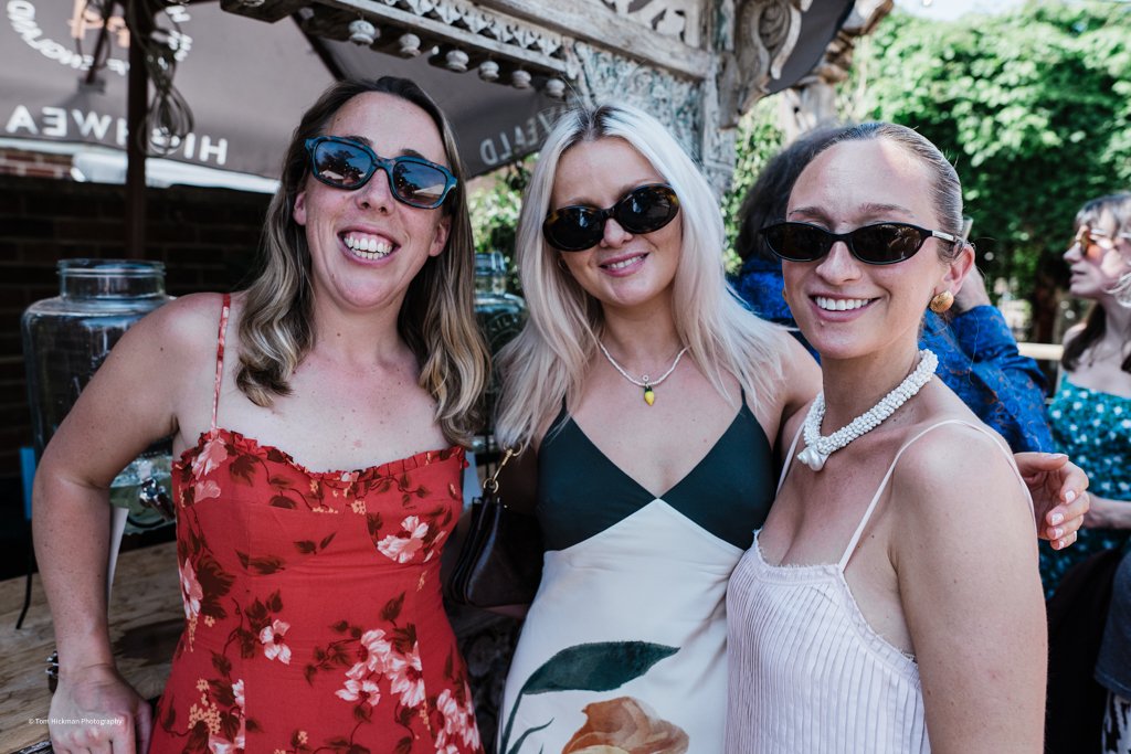 Three women smiling and posing for a photo at an outdoor event, wearing sunglasses and summer dresses.