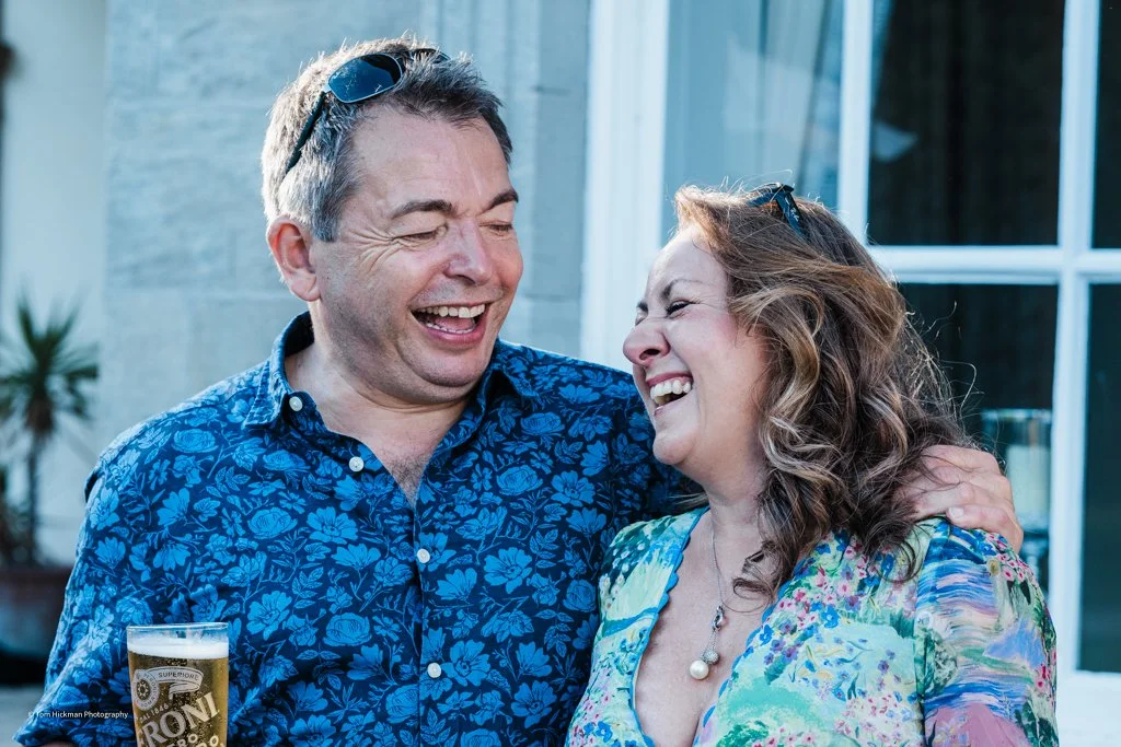 Two people, a man and a woman, are smiling and laughing together outdoors, with the man holding a glass of beer.