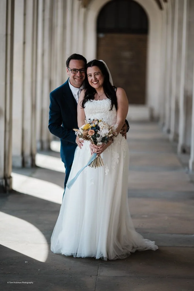 A bride and groom smiling and posing inside a rustic wooden corridor, with the bride holding a bouquet of flowers.