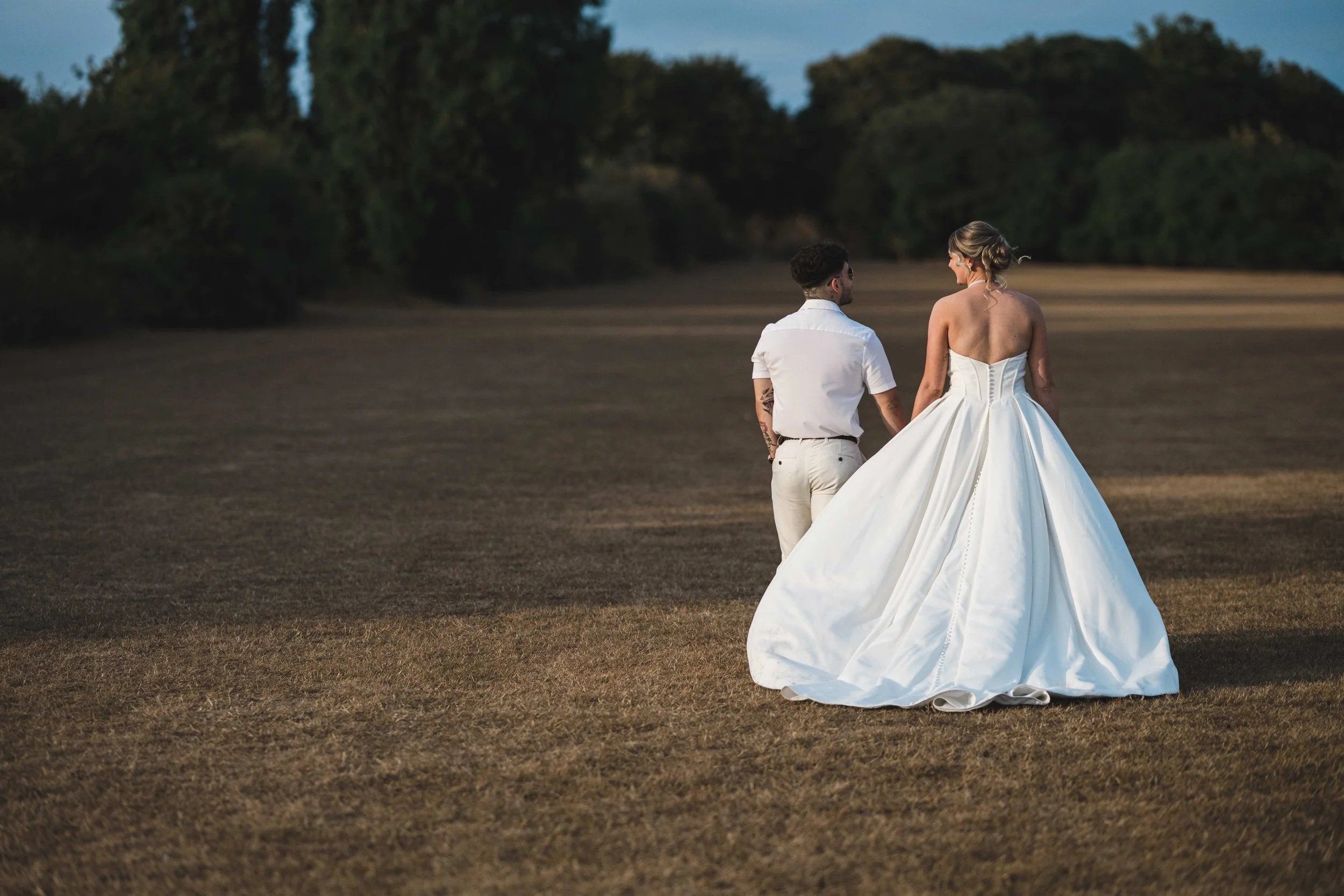 A couple dressed in wedding attire walking hand in hand across an open field with trees in the background during dusk.