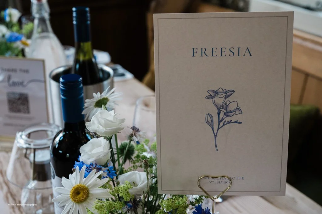 A wedding table centerpiece with a sign that says 'Freesia' and a floral illustration, alongside bottles of wine and a bouquet of daisies and other flowers.