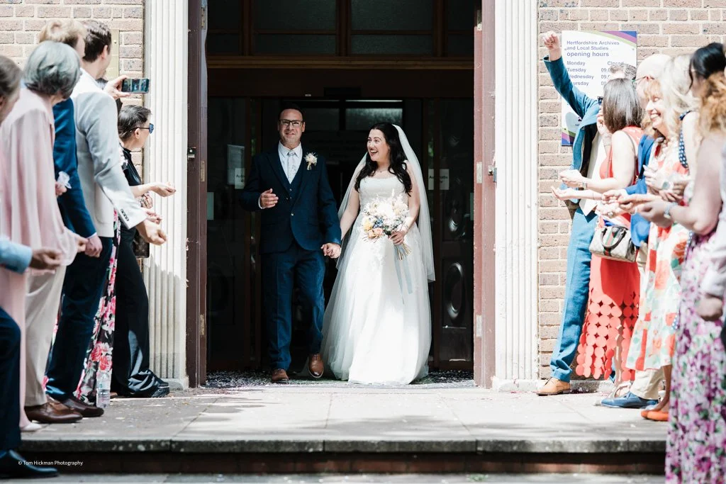 Bride and groom walking out of a building, smiling and holding hands, as friends and family cheer and take photos.