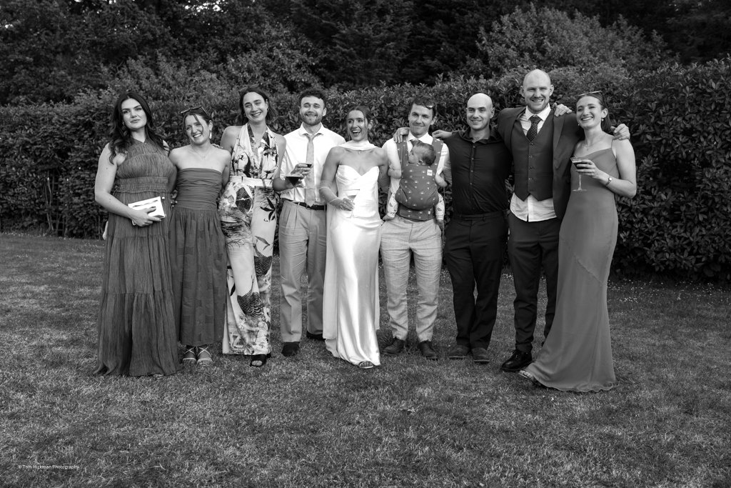 Group of people dressed in formal attire standing outdoors on grass with bushes in the background, smiling and posing for a photo.