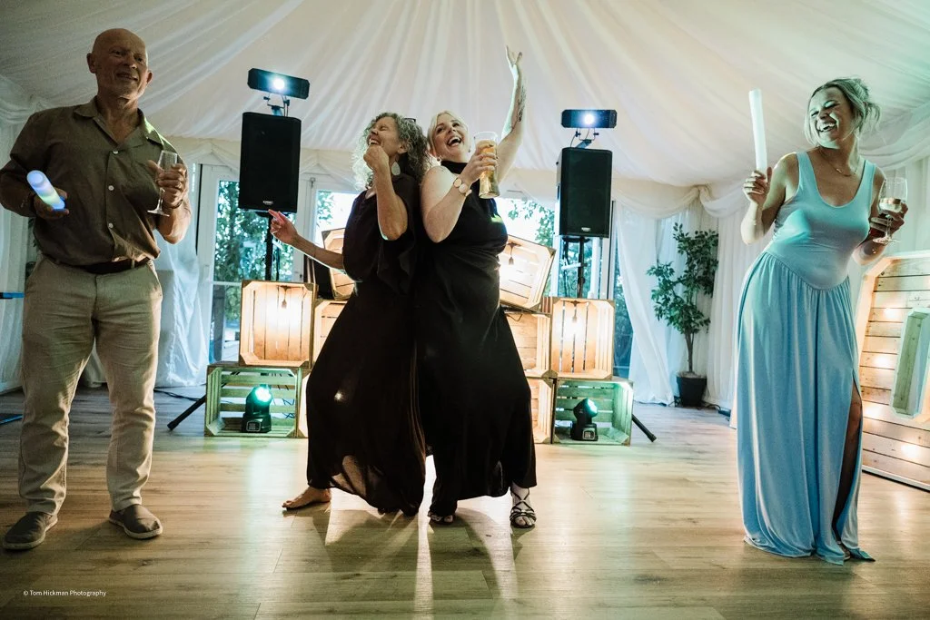 Four people dancing and celebrating at a party with drinks, in a decorated tent with lighting and plants in the background.