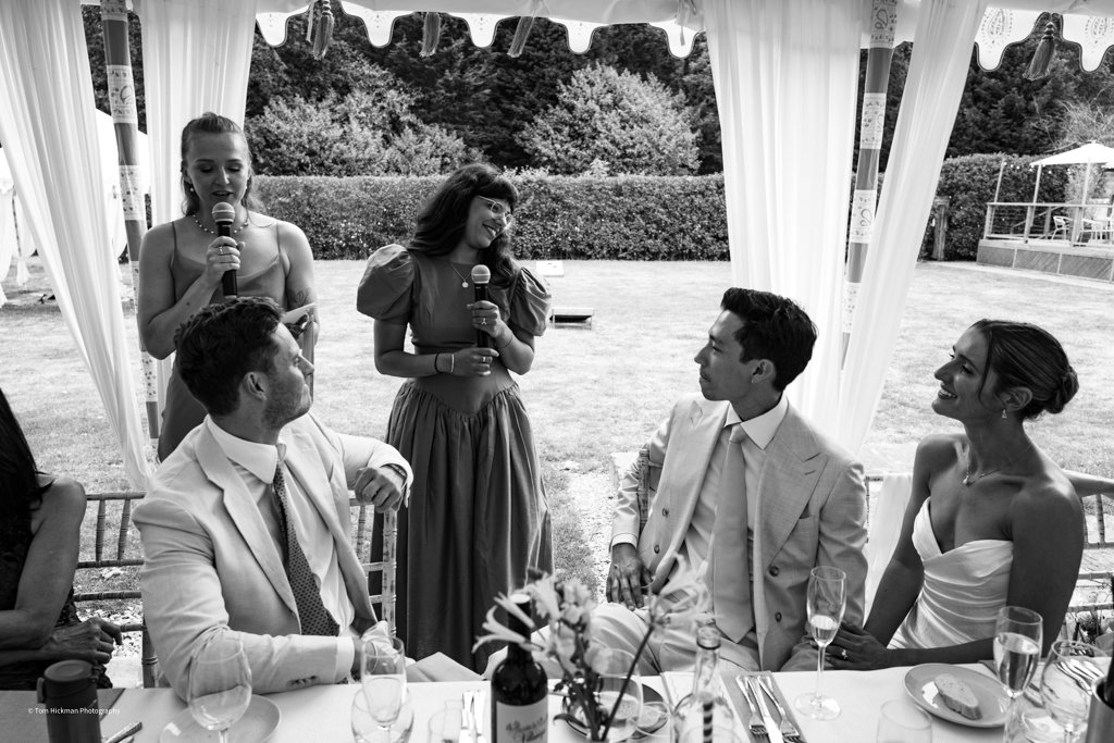 Black and white photo of a wedding reception inside a tent, with five people gathered around a table. Two women are standing and speaking into microphones, while three people are seated, engaged and listening. The outdoor background shows trees and a