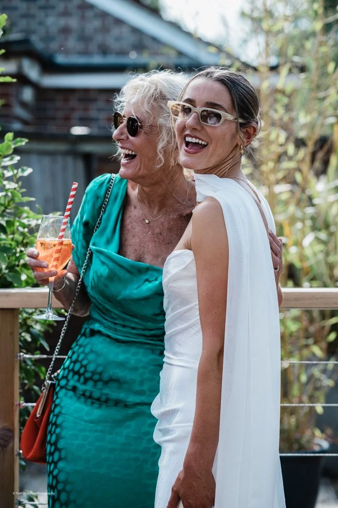Two women smiling and embracing outdoors, one holding a glass of orange beverage with a striped straw, surrounded by greenery.