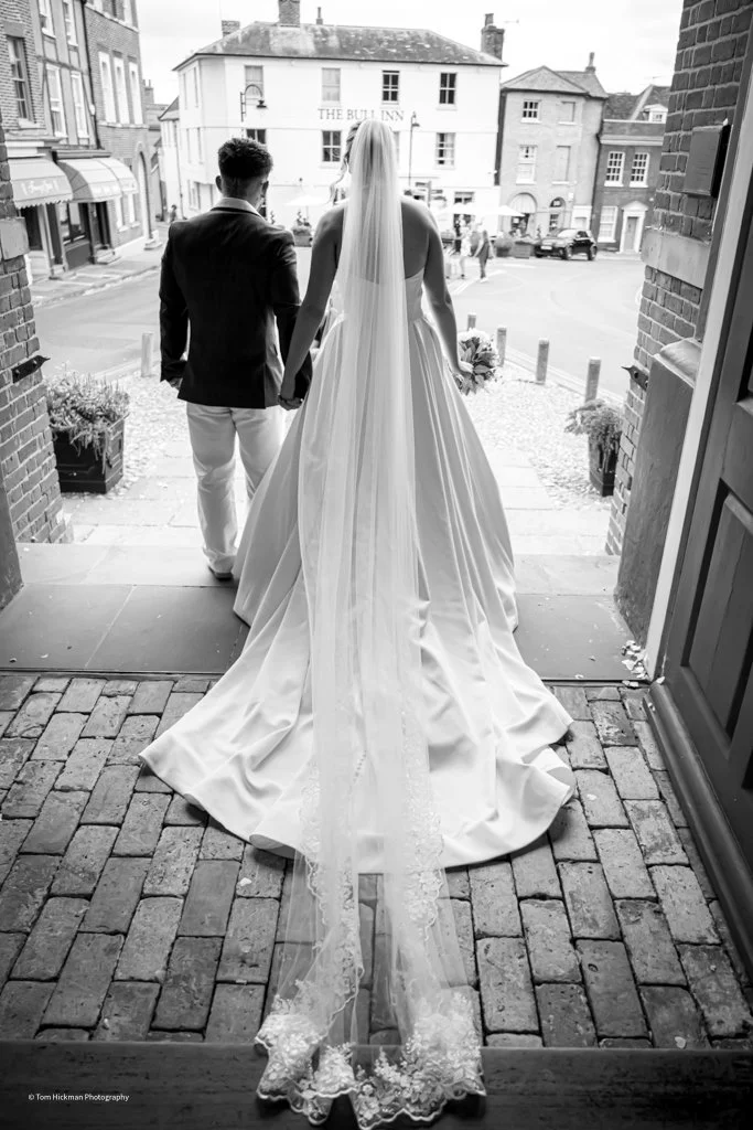 A bride and groom holding hands, walking out of a building onto a street, with the bride wearing a long wedding gown and veil, and the groom in a suit, captured in black and white.