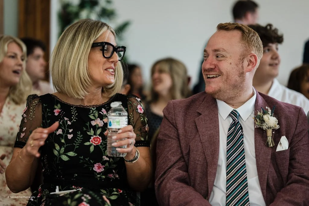 A woman with blonde hair, glasses, and a floral dress is smiling and holding a water bottle, engaging in conversation with a man in a maroon suit, white shirt, and striped tie, who is also smiling and wearing a small floral boutonniere, at a social g