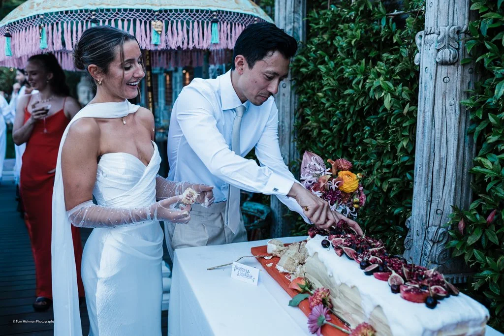 A wedding reception scene with a bride and groom cutting a large wedding cake decorated with berries, flowers, and whipped cream, outdoors with greenery and a decorative umbrella in the background.