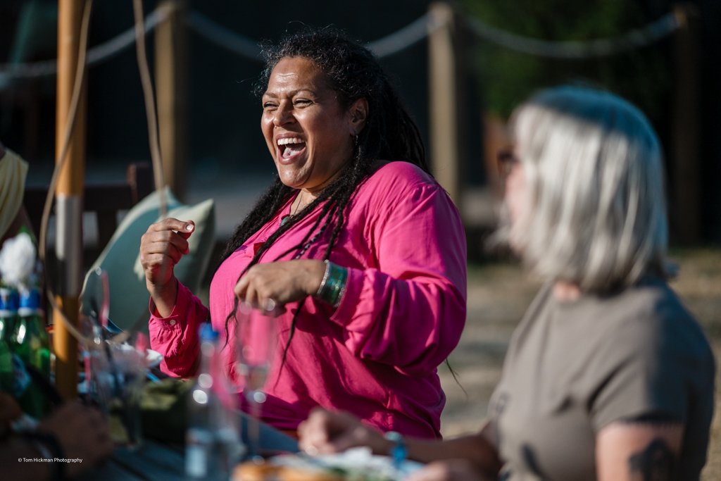 A woman with dreadlocks laughing and smiling while sitting at an outdoor gathering with others, wearing a bright pink jacket.