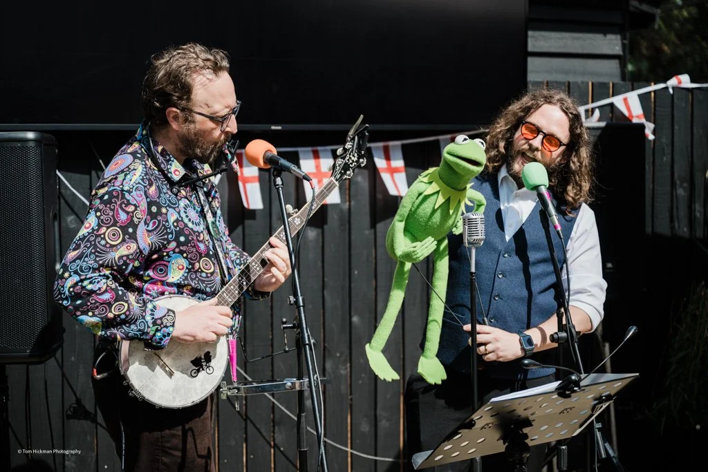 Two men performing with musical instruments and a puppet. The man on the left is playing a banjo, wearing a colorful patterned shirt, glasses, and has curly hair and a beard. The man on the right is holding a green puppet resembling Kermit the Frog, 
