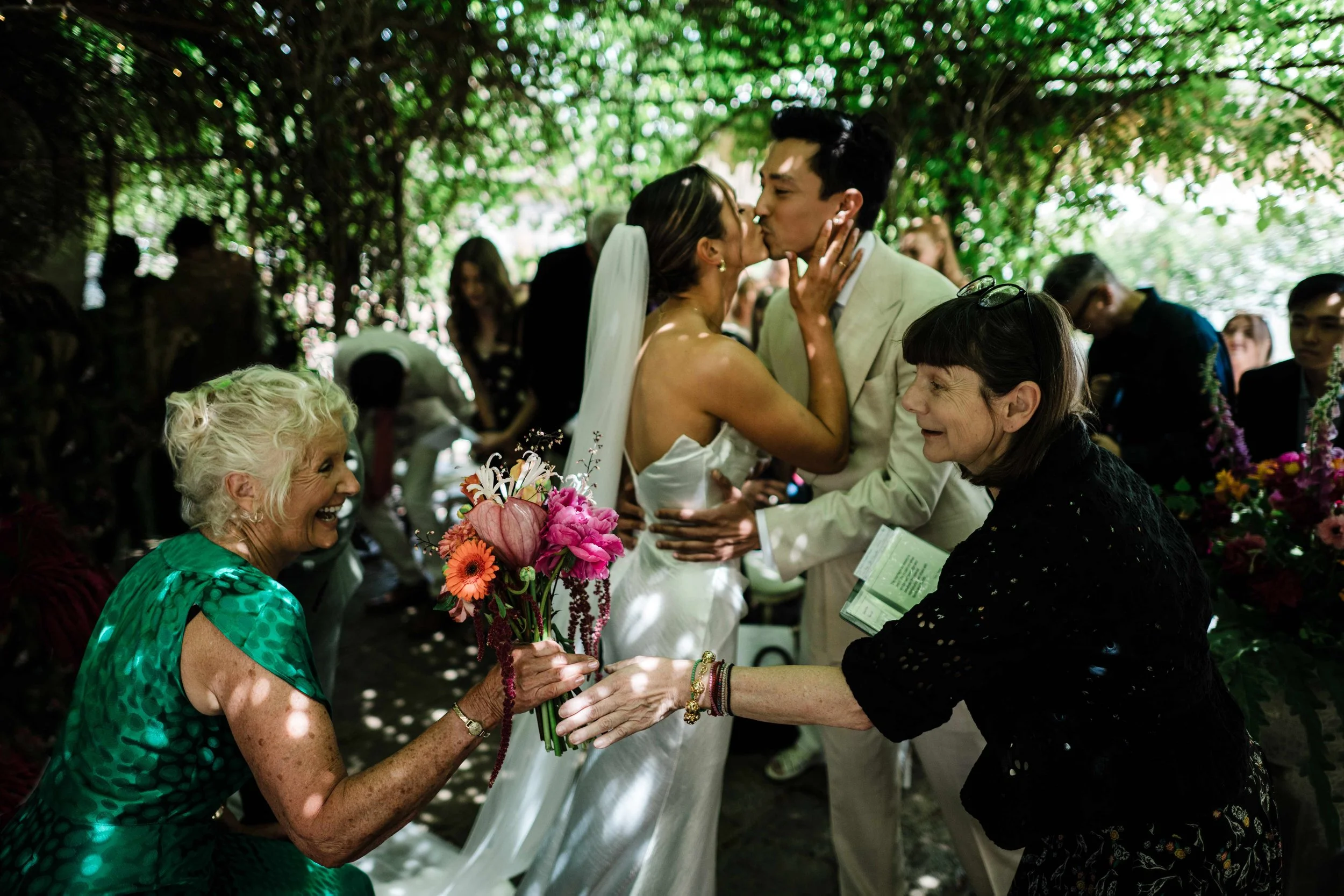 A bride and groom share a kiss at their wedding ceremony, surrounded by guests, under a canopy of green leaves during the daytime.