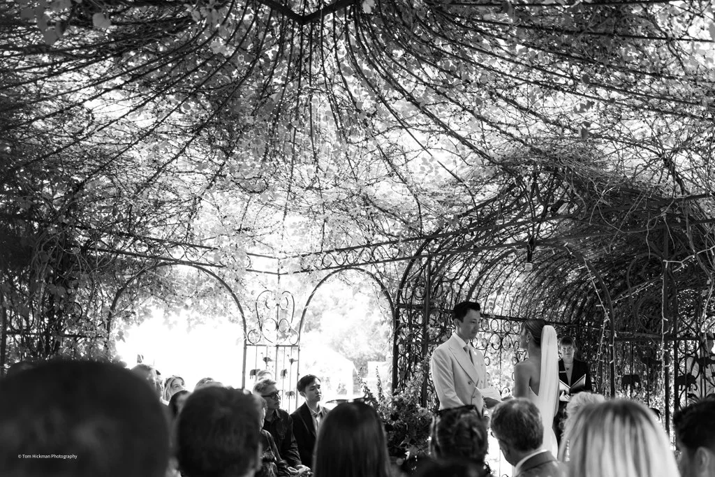 A black-and-white photo of a wedding ceremony taking place outdoors under an archway covered with trees.