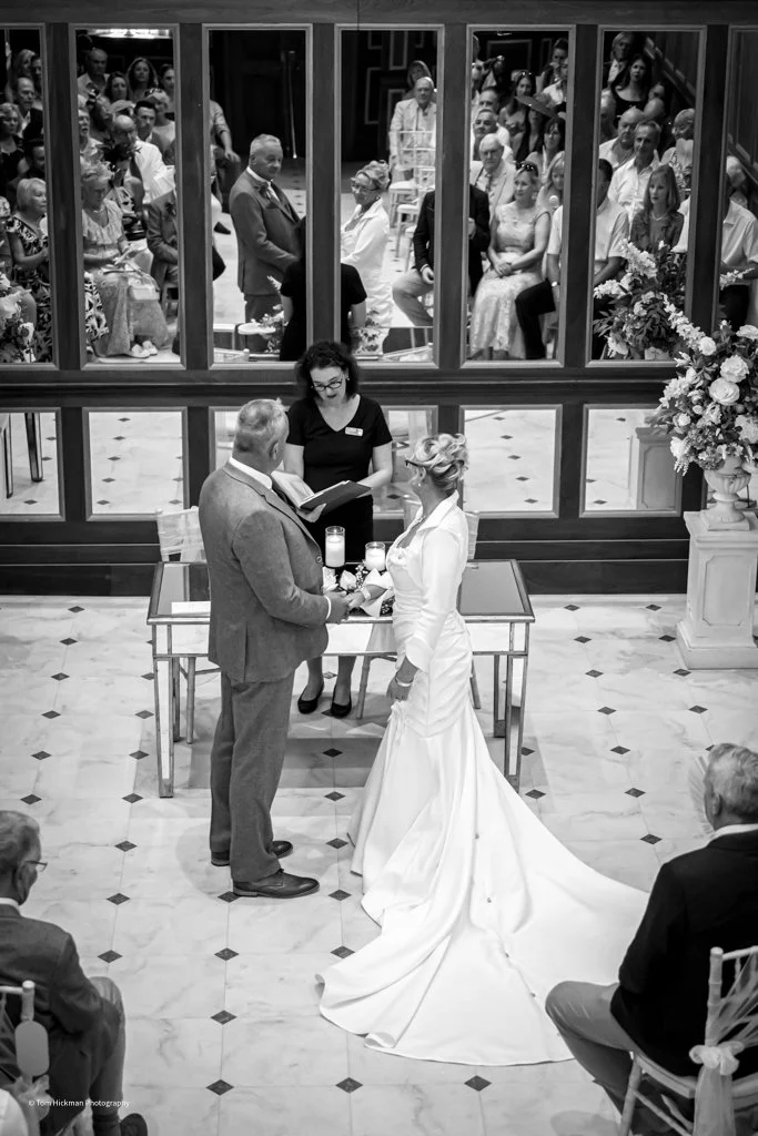 A wedding ceremony with a bride and groom exchanging vows, standing in front of an officiant. The bride is wearing a long white gown with a train, and the groom is dressed in a suit. Guests are seated behind a glass partition, watching the ceremony.