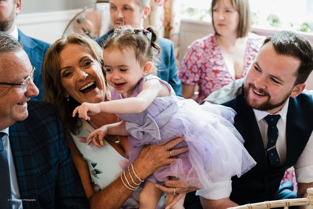 A group of people smiling and laughing, with a young girl in a purple dress being held up by an older woman, surrounded by adults at a celebration or gathering.