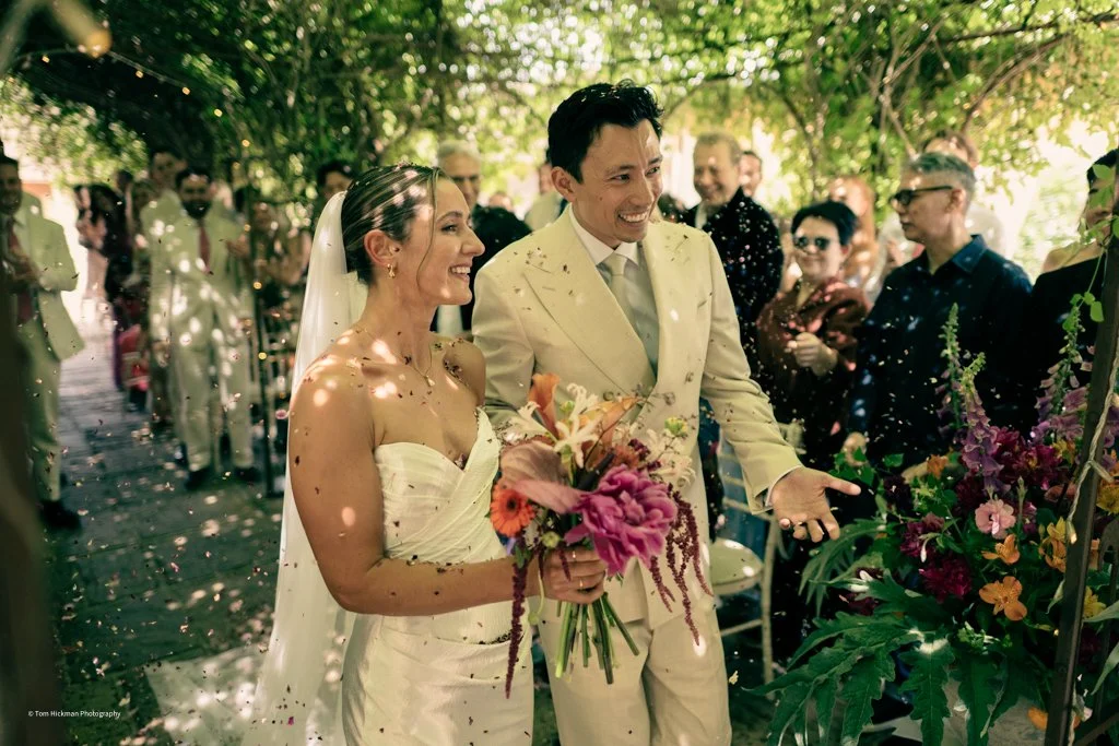 Bride and groom smiling during a wedding ceremony outside, surrounded by guests, with flowers and greenery.