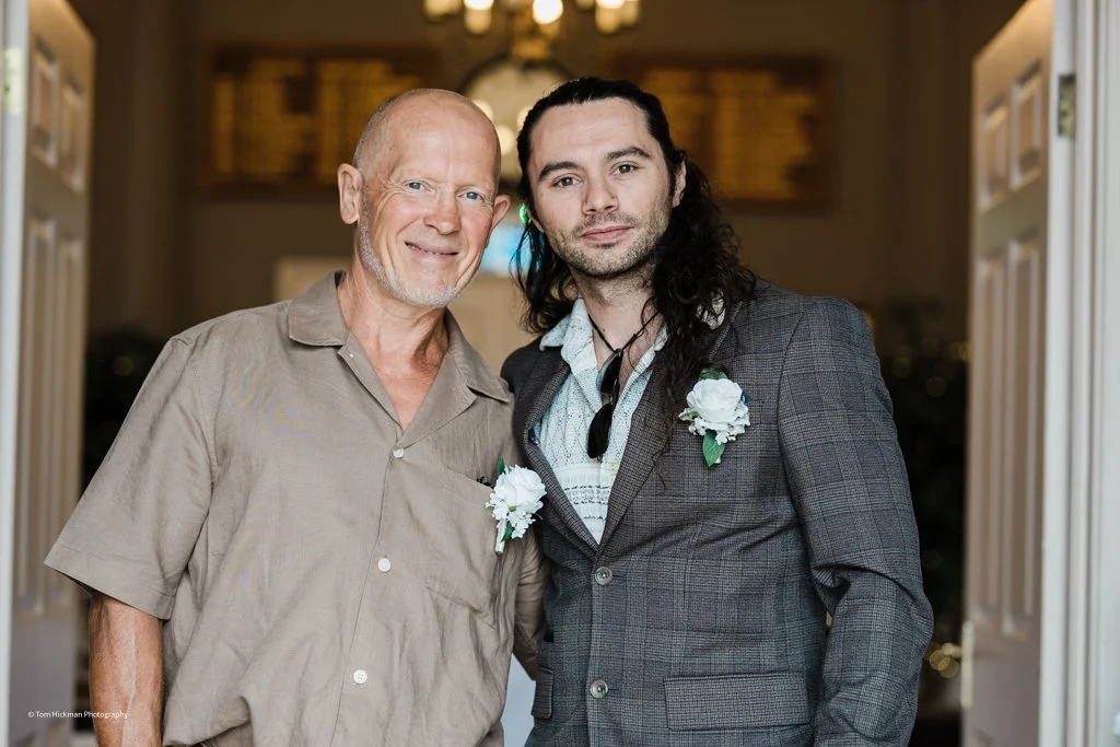 Two men stand together indoors, both wearing boutonnières, smiling at the camera. One is older with short, light hair, and the other is younger with long dark hair. They are dressed casually and formally, respectively.