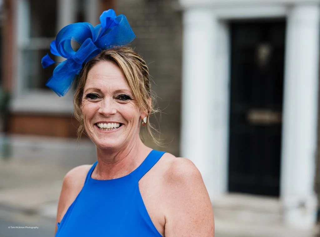 Smiling woman wearing a blue dress and large blue fascinator hat outdoors.