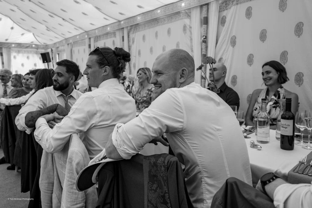 People sitting at a wedding reception table, smiling and socializing inside a decorated tent.
