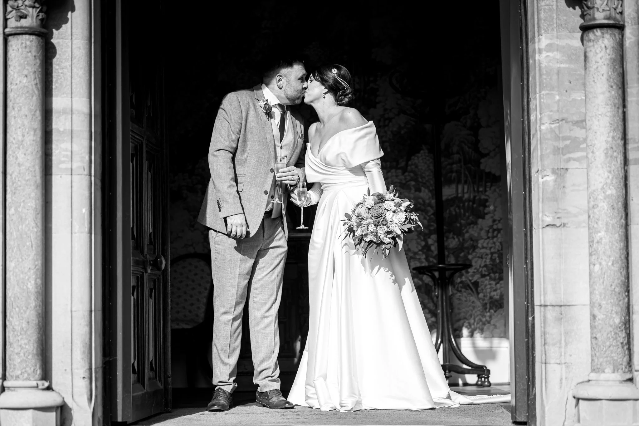 A black and white photograph of a bride and groom sharing a kiss at the entrance to Brownsover Hall Hotel during their wedding, with the groom holding a champagne glass and the bride holding a bouquet of flowers.