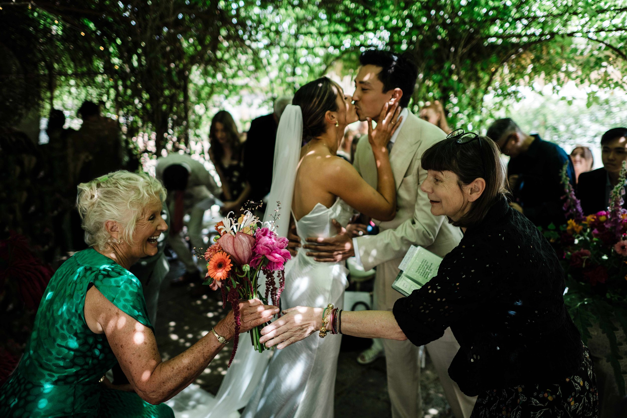 A wedding scene outdoors with a bride and groom kissing, surrounded by friends and family under a canopy of green leaves, with an elderly woman smiling and holding a bouquet of flowers.