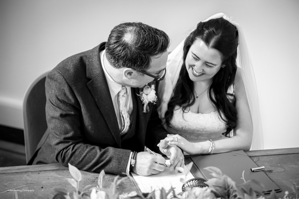 A bride and groom signing a wedding registry or marriage license at a table, smiling and holding hands.